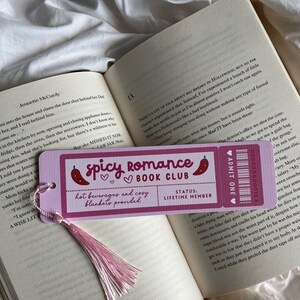 Pode incluir: Um marcador de livros rosa com o texto "Spicy Romance Book Club" e uma pimenta, em um livro aberto. O marcador de livros tem uma borla rosa e o texto "bebidas quentes e cobertores aconchegantes fornecidos."
