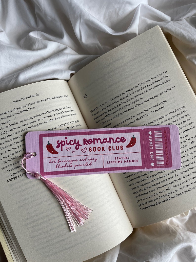 Pode incluir: Um marcador de livros rosa com o texto "Spicy Romance Book Club" e uma pimenta, em um livro aberto. O marcador de livros tem uma borla rosa e o texto "bebidas quentes e cobertores aconchegantes fornecidos."