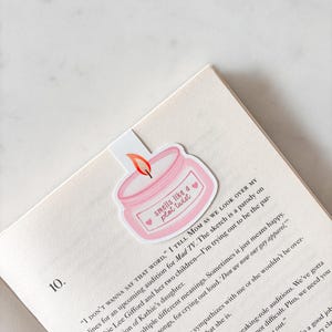 May include: A pink candle-shaped bookmark with the text "smells like a plot twist" and two hearts. The bookmark is placed on the corner of an open book with printed text. The bookmark has a flame.