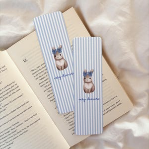 Può includere: Due segnalibri a righe blu e bianche con un'illustrazione ad acquerello di un coniglietto con un fiocco blu. Il testo "very demure" è scritto su ogni segnalibro. I segnalibri sono appoggiati su un libro aperto.