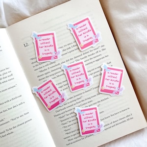 Könnte beinhalten: Aufgeschlagene Buchseiten mit mehreren rechteckigen, rosa Aufklebern. Jeder Aufkleber trägt den Text "a reader without her kindle is a tragedy". Die Aufkleber haben einen hellrosa Rand und florale Verzierungen.