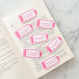 Può includere: Una collezione di adesivi rosa con la scritta "my social battery is running low" a forma di batteria, decorati con arcobaleni, lune e stelle. Gli adesivi sono sulle pagine di un libro aperto.