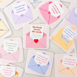 May include: A selection of square cards, each designed as an envelope with a reading prompt inside. The cards feature pastel colours and phrases like "Your Next Read is a Magical Realism Novel." The central card reads "the book lovers tbr cards."