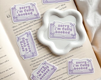 May include: A collection of lavender and white stickers with the text "sorry I'm fully booked" over an open book design. The stickers are arranged on an open book and a white decorative tray.
