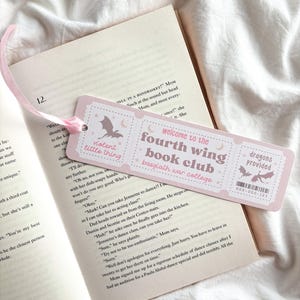 Op de afbeelding: Een roze boekenlegger met de tekst "violent little thing" en "welcome to the fourth wing book club basgiath war college." De boekenlegger heeft een vleermuis- en maan-afbeelding en een roze lint. De boekenlegger ligt op een open boek.