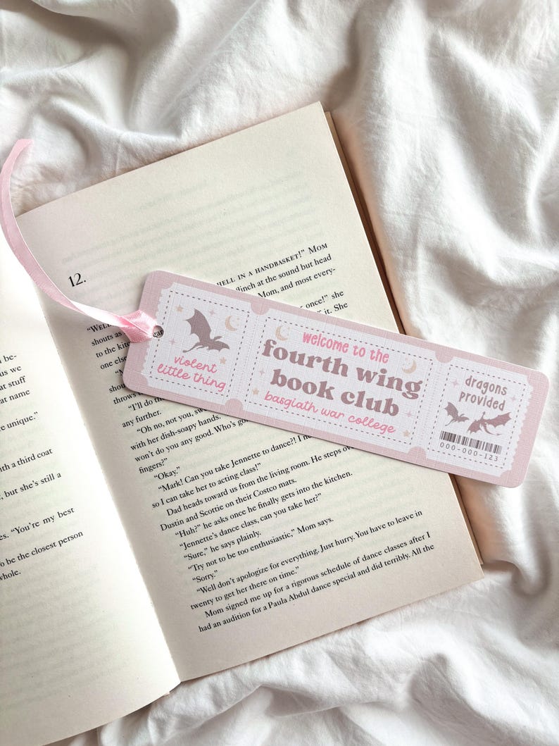 Op de afbeelding: Een roze boekenlegger met de tekst "violent little thing" en "welcome to the fourth wing book club basgiath war college." De boekenlegger heeft een vleermuis- en maan-afbeelding en een roze lint. De boekenlegger ligt op een open boek.