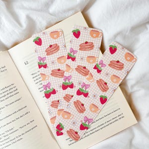 Puede incluir: Tres marcapáginas decorativos con un patrón repetido de fresas, panqueques, tazas de té y lazos sobre un fondo a cuadros rosa claro. Los marcapáginas están colocados sobre un libro abierto con texto.