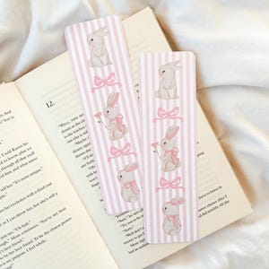 Può includere: Due segnalibri a righe rosa e bianche con illustrazioni di coniglietti e fiocchi rosa. I segnalibri sono appoggiati sulle pagine aperte di un libro. I coniglietti sono raffigurati in diverse pose, alcuni con fiori.