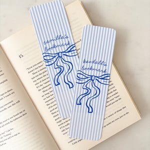May include: Two bookmarks with blue and white stripes, each bearing a name in cursive script: "camilla's bookmark" and "kendall's bookmark". A blue bow illustration adorns each bookmark. They are placed on an open book.