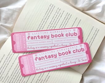 May include: Two pink "Fantasy Book Club" membership cards with white text, lying on an open book. The cards have a ticket design with the text "Fantasy is a necessary ingredient in living - Dr. Seuss."