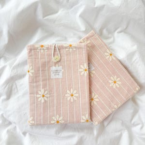 May include: Two pink fabric cases with white daisy embroidery and vertical stripes. One case is closed with a wooden button and string. The text "FLEURS ON SUNDAY" is printed on a small label. The cases are on a white sheet.