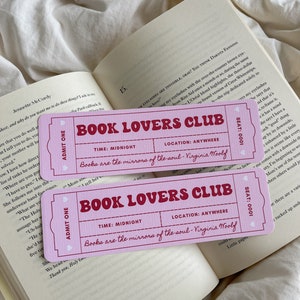 May include: Two pink ticket-shaped bookmarks with the text "BOOK LOVERS CLUB" in a bold, vintage font. The bookmarks are on top of an open book with white pages. The text "Books are the mirrors of the soul - Virginia Woolf" is also printed on the bookmarks.