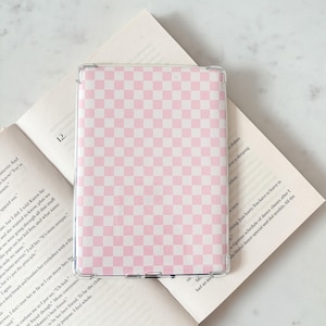 May include: A transparent phone case featuring a pink and white chequered design. The case is placed on an open book, with text visible on the pages. The book is open to page 12.