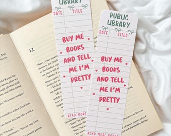 May include: Two rectangular bookmarks with a Public Library design, featuring spaces for date and title. The bookmarks have the text "BUY ME BOOKS AND TELL ME I'M PRETTY" in pink. One bookmark rests on an open book.