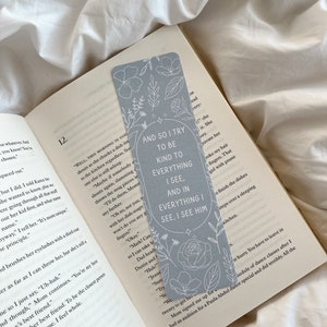 Puede incluir: Un marcap&aacute;ginas azul claro con dise&ntilde;os florales y el texto "AND SO I TRY TO BE KIND TO EVERYTHING I SEE, AND IN EVERYTHING I SEE, I SEE HIM." El marcap&aacute;ginas est&aacute; en un libro abierto.