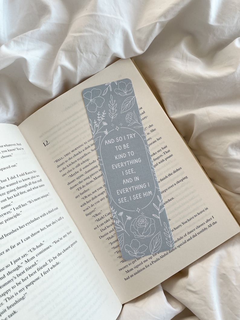 Puede incluir: Un marcap&aacute;ginas azul claro con dise&ntilde;os florales y el texto "AND SO I TRY TO BE KIND TO EVERYTHING I SEE, AND IN EVERYTHING I SEE, I SEE HIM." El marcap&aacute;ginas est&aacute; en un libro abierto.