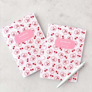 May include: Two notebooks with a cherry pattern and a white pen. The notebooks have a white background with a repeating pattern of red cherries and pink heart-shaped designs. Each notebook has a pink label that reads "NOTEBOOK" and "32 lined pages".