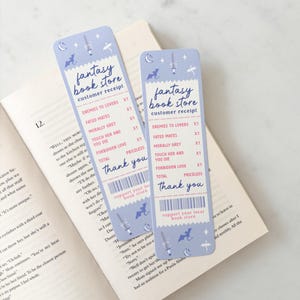 Op de afbeelding: Twee lichtblauwe fantasy-thema bladwijzers die op bonnen lijken, met de tekst "Fantasy Book Store" en titels van boeken. De bladwijzers bevatten illustraties van een zwaard, draak en maan.