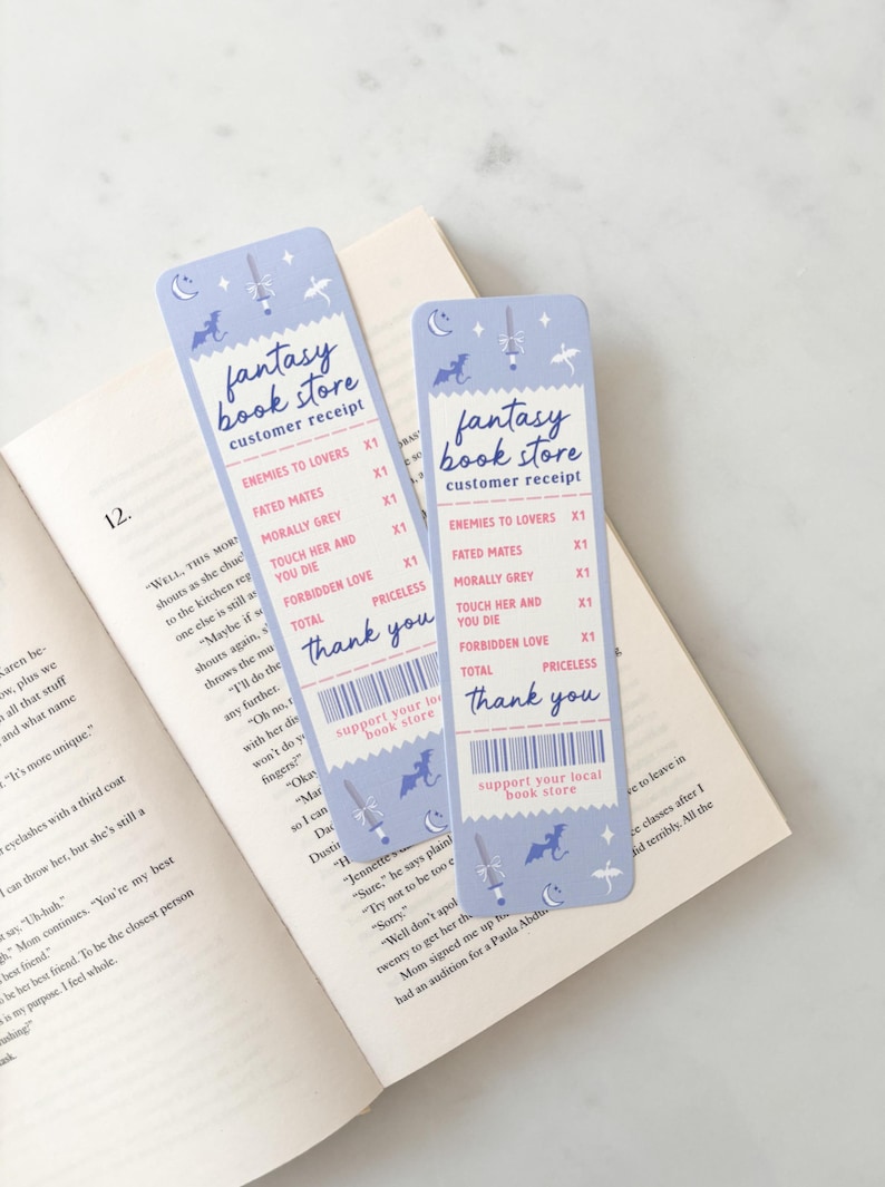 May include: Two light blue fantasy-themed bookmarks, designed like receipts, with "Fantasy Book Store" and book titles. They feature illustrations of a sword, dragon, and moon. The bookmarks are on a book.