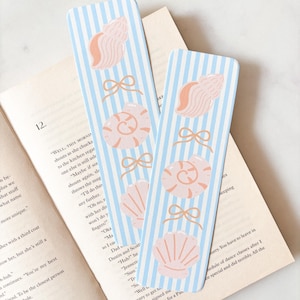 May include: Two rectangular bookmarks featuring a light blue and white vertical striped pattern. Each bookmark is decorated with illustrations of three pink seashells and three small tan bows arranged in an alternating vertical sequence.