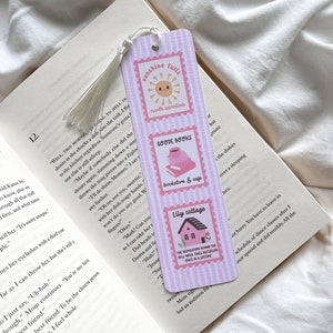 May include: A pink and white striped bookmark with three rectangular stamp designs. The top stamp reads "Sunshine Falls, North Carolina" with a smiling sun. The middle stamp says "Goode Books, bookstore & cafe". The bottom stamp says "Lily cottage".