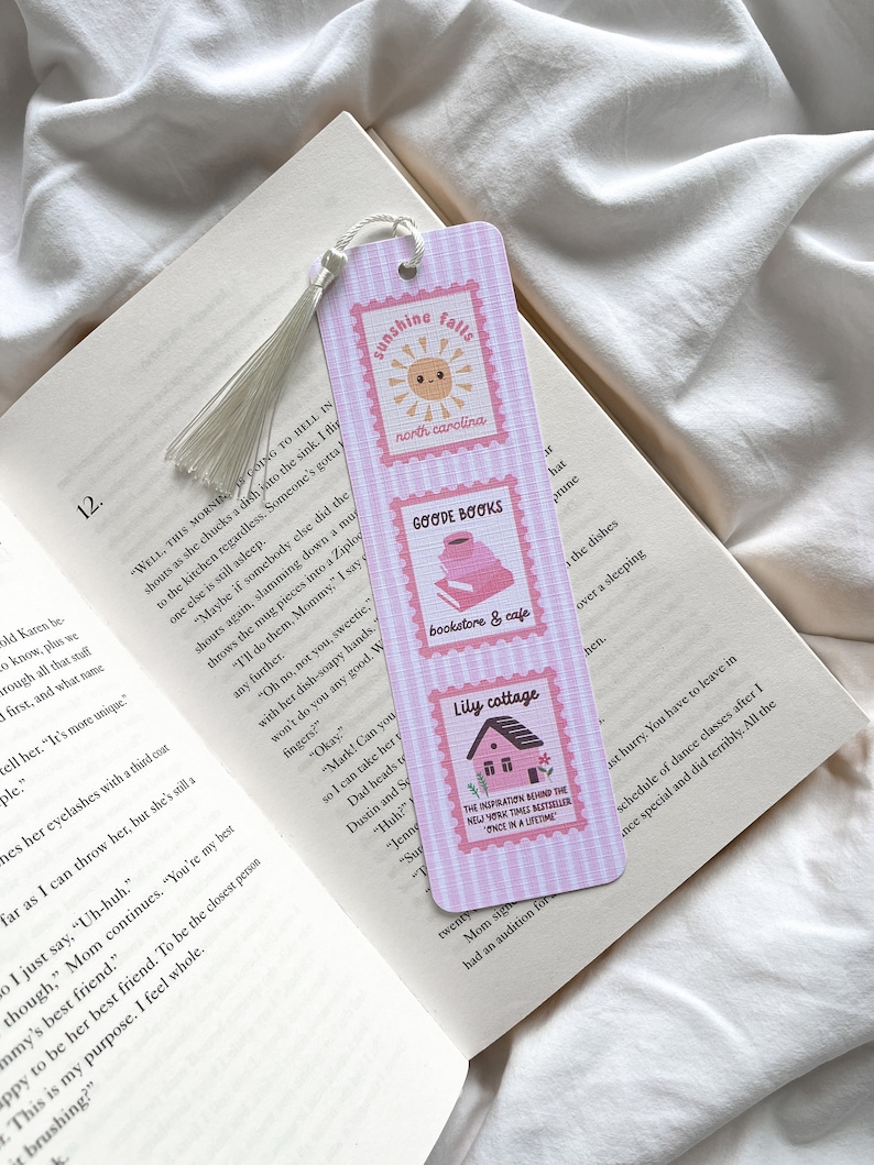 May include: A pink and white striped bookmark with three rectangular stamp designs. The top stamp reads "Sunshine Falls, North Carolina" with a smiling sun. The middle stamp says "Goode Books, bookstore & cafe". The bottom stamp says "Lily cottage".