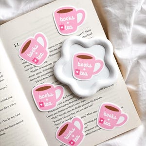 May include: Pink stickers in the shape of a teacup with the words "books + tea" printed on them. The teacups have a heart-shaped tea bag tag. The stickers are scattered on an open book.