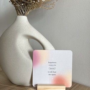 Puede incluir: Una tarjeta cuadrada con un degradado rosa y naranja, sujeta por un peque&ntilde;o bloque de madera. La tarjeta tiene el texto "happiness comes in waves it will find me again". Un jarr&oacute;n blanco y flores secas est&aacute;n en el fondo.
