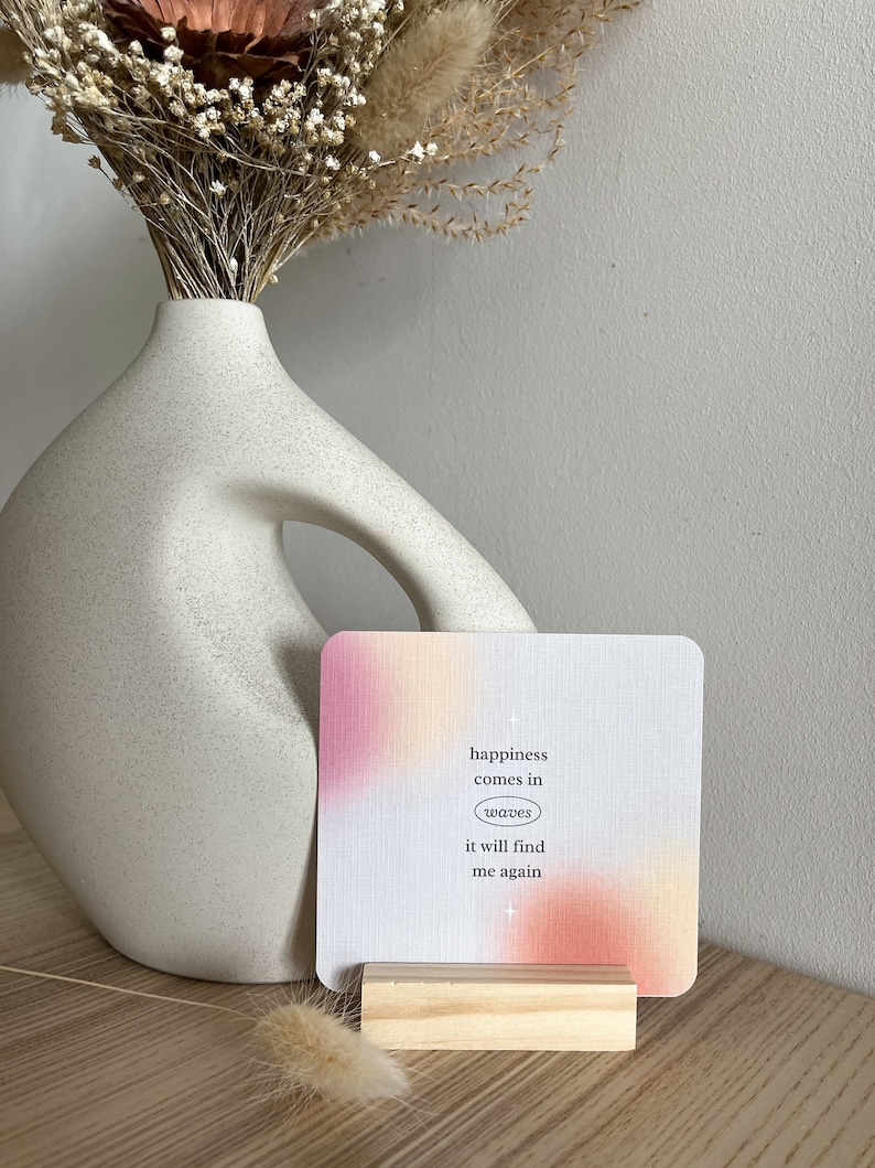 Puede incluir: Una tarjeta cuadrada con un degradado rosa y naranja, sujeta por un peque&ntilde;o bloque de madera. La tarjeta tiene el texto "happiness comes in waves it will find me again". Un jarr&oacute;n blanco y flores secas est&aacute;n en el fondo.
