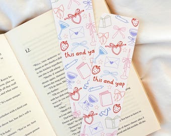 May include: Two decorative bookmarks with a repeating pattern of bows, hearts, strawberries, and other small illustrations. The text "this and ya" is printed on the bookmarks. The bookmarks are placed on top of an open book.