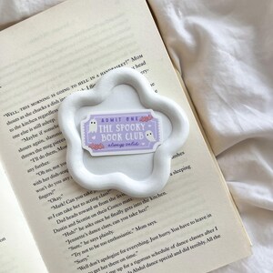 May include: A white, cloud-shaped ceramic dish holds a sticker that reads "ADMIT ONE THE SPOOKY BOOK CLUB always valid." The sticker features a ghost and bats. The dish rests on an open book with printed text.