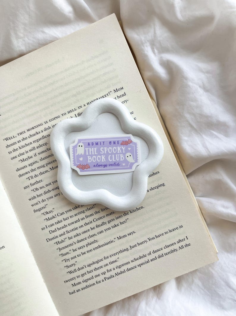 May include: A white, cloud-shaped ceramic dish holds a sticker that reads "ADMIT ONE THE SPOOKY BOOK CLUB always valid." The sticker features a ghost and bats. The dish rests on an open book with printed text.