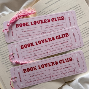 May include: Three pink bookmarks with the text "BOOK LOVERS CLUB" in red. Each bookmark has a pink tassel and the quote "Books are the mirrors of the soul - Virginia Woolf." The bookmarks are on top of an open book.