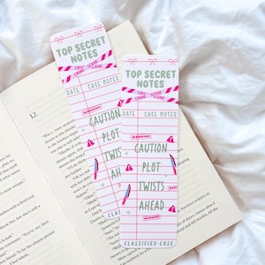 Puede incluir: Dos marcadores rectangulares con un diseño de lazo en la parte superior, con el texto "TOP SECRET NOTES". Los marcadores tienen una combinación de colores rosa y blanco con gráficos de cinta de escena del crimen y las palabras "CAUTION", "PLOT" y "TWISTS AHEAD".