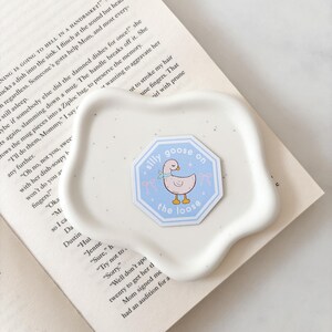 Op de afbeelding: Een lichtblauwe sticker met de tekst "silly goose on the loose" en een cartoon-gans-illustratie. De sticker ligt op een wit, organisch gevormd schaaltje, dat op een open boek ligt.