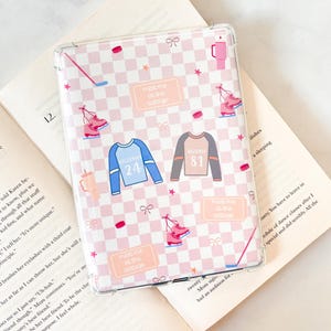 May include: Transparent phone case with a pink and white chequered design. The case has illustrations of ice skates, hockey sticks, and jerseys with the numbers 24 and 81. Text on the case reads "meet me at the cottage."
