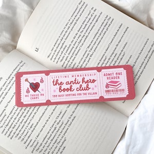Może przedstawiać: Różowa zakładka do książki z napisem "Lifetime Membership The Anti Hero Book Club" i "Admit One Reader" na otwartej książce. Zakładka ma serce ze strzałą i słowa "We Thrive on Chaos."