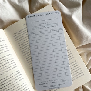 K&ouml;nnte beinhalten: Eine graue Bibliothekskarte mit dem Text "FROM THE LIBRARY OF:" und einem Raster zur Aufzeichnung von Buchdetails. Die Karte liegt auf einem offenen Buch mit sichtbarem Text.