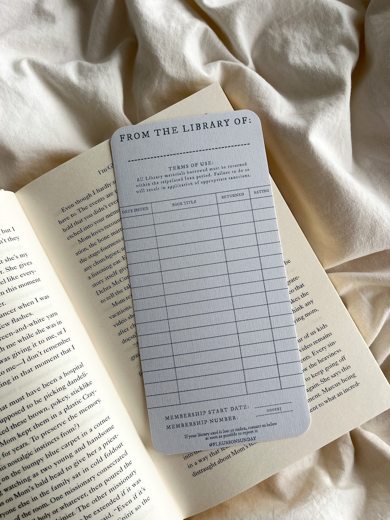 K&ouml;nnte beinhalten: Eine graue Bibliothekskarte mit dem Text "FROM THE LIBRARY OF:" und einem Raster zur Aufzeichnung von Buchdetails. Die Karte liegt auf einem offenen Buch mit sichtbarem Text.