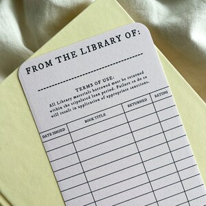 K&ouml;nnte beinhalten: Ein wei&szlig;es Bibliotheksk&auml;rtchen mit schwarzem Text, darunter "FROM THE LIBRARY OF:" und "TERMS OF USE." Die Karte liegt auf einem hellgelben Buch. Die Karte hat Felder f&uuml;r Buchtitel, Ausgabedatum und Bewertung.