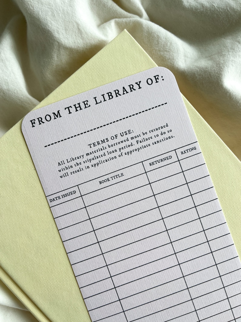K&ouml;nnte beinhalten: Ein wei&szlig;es Bibliotheksk&auml;rtchen mit schwarzem Text, darunter "FROM THE LIBRARY OF:" und "TERMS OF USE." Die Karte liegt auf einem hellgelben Buch. Die Karte hat Felder f&uuml;r Buchtitel, Ausgabedatum und Bewertung.