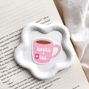 May include: A white, flower-shaped ceramic coaster holds a pink sticker with a teacup design. The teacup has the words "books + tea" and a heart on it. The coaster rests on the open pages of a book.