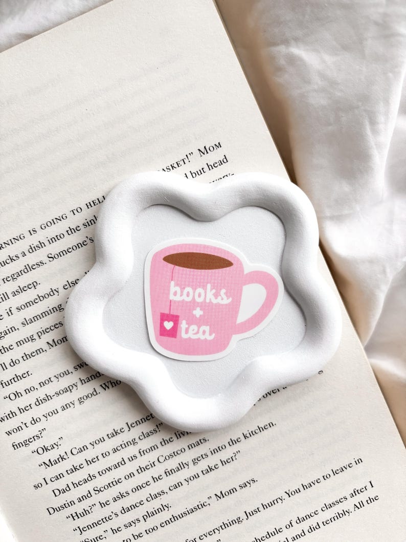 May include: A white, flower-shaped ceramic coaster holds a pink sticker with a teacup design. The teacup has the words "books + tea" and a heart on it. The coaster rests on the open pages of a book.