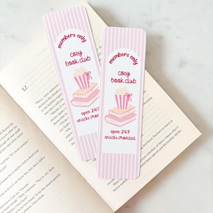 Peut inclure: Deux marque-pages à rayures roses et blanches avec le texte "members only cosy book club" et "open 24/7 snacks provided". Chaque marque-page présente un dessin de livres et de pop-corn. Les marque-pages reposent sur un livre ouvert.