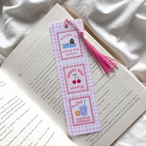 May include: A pink and white gingham bookmark with a pink tassel. The bookmark features three postage stamp-style designs with the text "WELCOME TO waning bay, michigan", "CHERRY HILL winery", and "miles LAVENDER LEMONADE". The bookmark is placed in an open book.