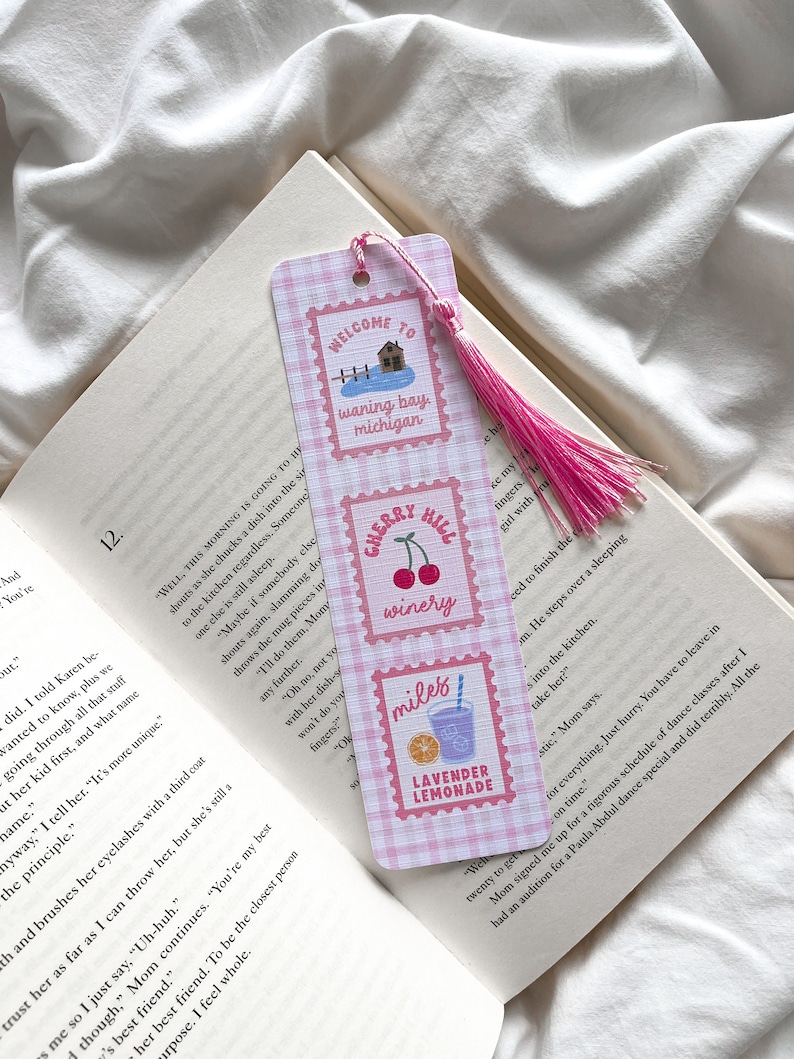 May include: A pink and white gingham bookmark with a pink tassel. The bookmark features three postage stamp-style designs with the text "WELCOME TO waning bay, michigan", "CHERRY HILL winery", and "miles LAVENDER LEMONADE". The bookmark is placed in an open book.