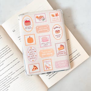 以下が含まれることがあります： グリッドパターンと様々なステッカーデザインが施された透明な携帯電話ケース。ステッカーには、リンゴ、カボチャ、「I am an autumn」、「Luke's pancake stack」などの秋をテーマにしたイラストが描かれています。