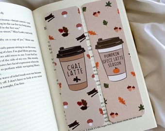 May include: Two beige bookmarks with fall-themed illustrations are placed in an open book. One bookmark features a Chai Latte design, and the other has a Pumpkin Spice Latte design. Both have autumn leaves, mushrooms, and acorns.
