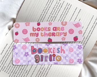 May include: Two rectangular bookmarks with text and decorative elements. The top bookmark reads "books are my therapy" with smiley faces, flowers, and strawberries. The bottom bookmark says "bookish girlie" with a checkered pattern.