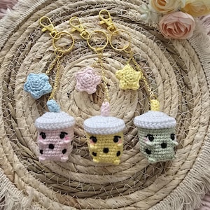 May include: Three crochet boba tea keychains in pink, yellow, and green, each with a white lid and gold-colored hardware. The keychains are adorned with crochet star charms in blue, pink, and yellow. The keychains are displayed on a woven mat.